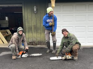 A photo of three men with chainsaws outside of a barn.